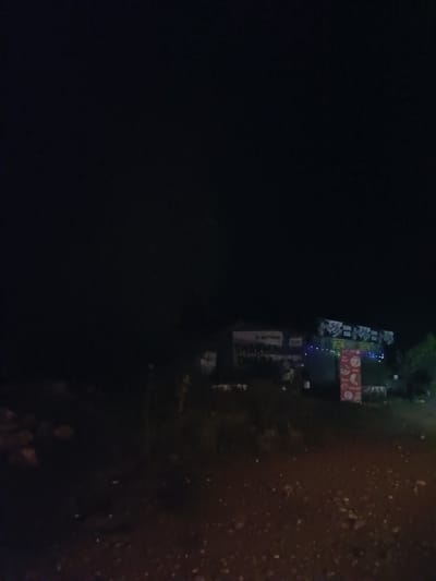 The image shows a nighttime scene in Alsu, India, featuring several illuminated signs and a dimly lit environment. In the foreground, a gravelly, uneven surface is visible, suggesting a rural or undeveloped area. Several structures, possibly shops or roadside stalls, are present in the background. One prominent sign displays text that appears to be in Hindi, with some words partially obscured by darkness and lighting. Another sign, red and rectangular, features circular graphics, likely advertisements. Colorful string lights adorn one of the structures, adding a festive touch. The overall impression is of a roadside establishment operating at night, possibly a food stall or a small business. The darkness and limited visibility suggest the image was taken in a location with minimal street lighting.