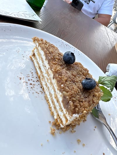 A slice of layered cake, topped with blueberries and a mint leaf, rests on a white plate. The cake has thin, golden-brown layers interspersed with white cream and a crumbly, nut-based coating on the exterior. Crumbles of cake are scattered on the plate. A fork is positioned to the right of the plate, with its tines partially visible. In the background, a wooden table and a white napkin are visible. A person in a white t-shirt, holding a phone, is also in the background, as is a green bottle and a white paper with a drawing on it. The scene appears to be an outdoor setting, possibly a cafe or restaurant, in Shkorpilovtsi, Bulgaria.