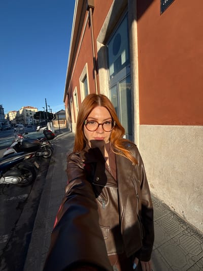 A selfie portrait shows a woman with red hair and glasses standing on a sidewalk in Sanremo, Italy. She is wearing a brown leather jacket. The background includes buildings, motorcycles parked along the curb, and a clear blue sky. The photo appears to have been taken during the day with sunlight illuminating the scene. The tiled sidewalk suggests an urban setting.