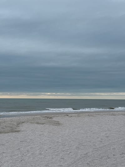 The image displays an uninhabited beach scene in Naples, United States of America, under an overcast sky. The upper two-fifths of the frame is occupied by a dense, uniform layer of dark grey clouds. A narrow horizontal band of pale yellow-grey light is visible at the horizon, situated between the cloud cover and the ocean. The middle section of the image features the ocean, characterized by dark grey-green water, with multiple small waves capped by white foam approaching the shore. The lower two-fifths of the frame shows an expansive area of light grey-white sand beach. No individuals, animals, or man-made objects are visible, and no specific actions or interactions are depicted within the scene.