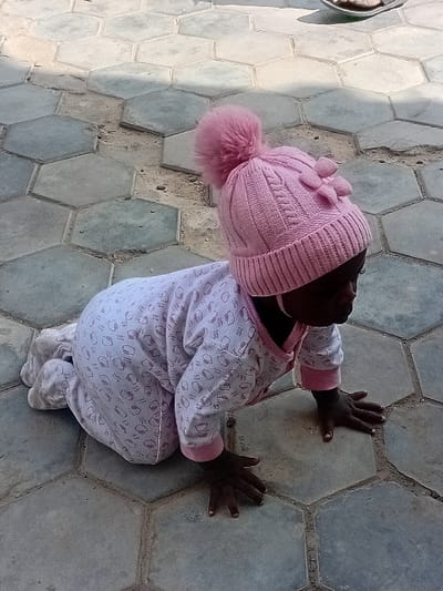 An infant is positioned on a paved surface in a crawling posture, resting on hands and knees, within the city of Kwazailewa, Nigeria. The infant, with dark skin, wears a light pink long-sleeved romper adorned with white patterns and pink trim at the collar and cuffs. A knitted pink beanie hat with a large pink pom-pom on top and a smaller decorative element on the right side covers the head. The infant's head is turned slightly to the right, looking downwards. The ground consists of unevenly laid hexagonal concrete paving tiles, exhibiting variations in shade from light to dark gray, with visible dirt and gaps between some tiles. Ambient light casts subtle shadows on the tiled surface. A blurry object, possibly a sandal or a dish, is partially visible in the upper right background.