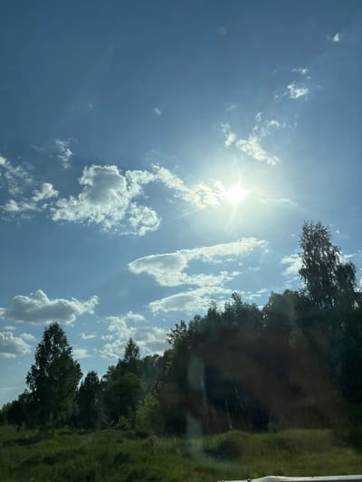 The image shows a bright, sunny sky with scattered white clouds. The sun is high in the sky, creating a strong glare. Along the top right edge of the image, there are trees and greenery. The image appears to be taken from a low angle, possibly from inside a car, looking up at the sky and trees. The location is Kvarsa, Russia.