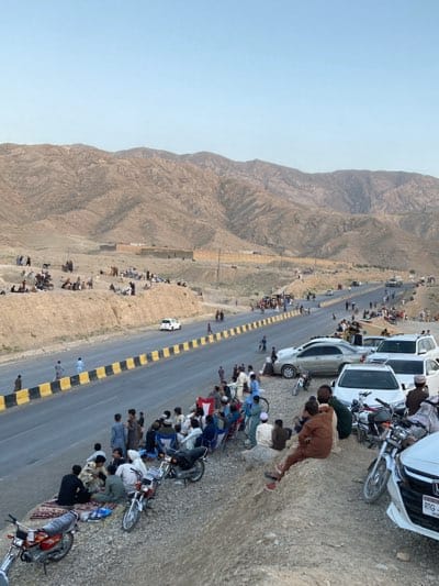 The image depicts a roadside scene in Kuchlak, Pakistan. A two-lane road bordered by a black and yellow barrier divides the foreground. On one side of the road, people and cars are present. There is a group of men seated on the embankment alongside motorcycles. Further along the road are more parked cars, people standing, and a few walking across the road. In the background, arid mountains rise against a light sky. Additional individuals are scattered across the landscape at the foot of the mountains. A building with a walled perimeter is visible in the distance.