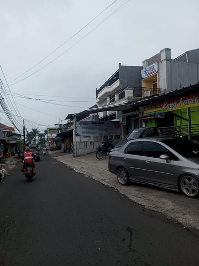 The image captures an overcast daytime street scene in the city of Houaythat, Laos. A paved asphalt road, displaying minor cracking, extends into the distance, curving slightly to the left.
In the foreground, a person wearing a red shirt is operating a dark-colored scooter with license plate "2553 KA," driving away from the viewer in the left lane. Further down the road, a dark car and another scooter are visible. The left side of the street features multiple utility poles with a dense entanglement of black wires, and a series of single and multi-story buildings, some appearing residential, recede into the background. Palm trees are visible further down the street on the left.
On the right side of the road, a silver sedan with multi-spoke custom rims is parked on the concrete pavement, facing away from the viewer. Behind it, a black pickup truck is parked, partially obscuring a blue motorcycle. A multi-story concrete building dominates the right side, featuring a yellow-green sign reading "Warung Nasi Kang Faris" on the ground floor. Above it, on the second floor, a white and blue sign identifies "Sagara Medika Cipanas PRAKTEK DOKTER 24 JAM" (Doctor's Practice 24 Hours). The building exhibits balconies with dark railings.
The sky is uniformly light grey and overcast. Numerous black utility wires are strung across the upper portion of the frame, with a line of small birds perched on one of the lower horizontal wires.
