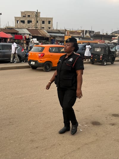 A police officer in full uniform stands on a street in what appears to be a busy market area. She wears a black uniform, including a cap, and is positioned near several parked cars, including an orange hatchback. Other vehicles, like tuk-tuks, are visible in the background. Buildings and stalls line the street, indicating a commercial setting. The sky is overcast, providing soft lighting for the scene. The police officer's presence suggests she might be on duty, monitoring the area.