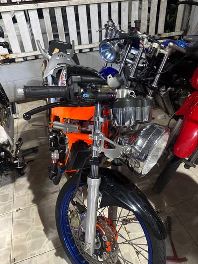 The image captures a close-up, eye-level view of several motorcycles parked indoors, possibly in a garage or workshop, in Si Sunthon, Thailand. The primary focus is on a bright orange motorcycle frame with a black fender and a prominent chrome headlight assembly. The motorcycle has a blue rim on its front wheel, adorned with a disc brake and purple bolts. A silver steering damper is visible on the frame, along with black handlebars. To the left, parts of another motorcycle are visible, including a silver fairing and black grips. Further in the background, a blue motorcycle with a round headlight and a red motorcycle with a red fender are partially visible. The environment appears to be dimly lit, with a tiled floor and a white slatted wall in the background.