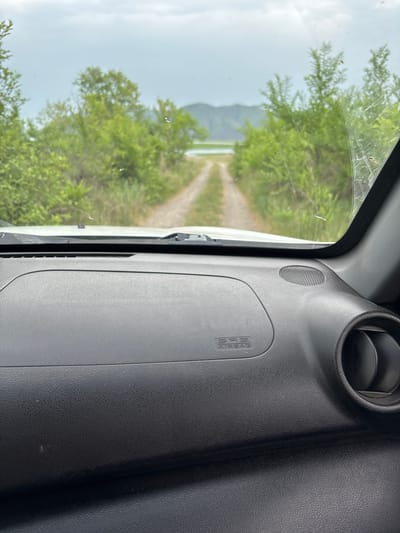 The image shows a view from inside a vehicle, looking out through the windshield. A dirt road leads into the distance, flanked by lush green trees and vegetation. In the background, a body of water is visible, with hills beyond it under a cloudy sky. The dashboard of the car is in the foreground, with an "SRS AIRBAG" label visible on the passenger side. The scene appears to be in a rural or natural setting, possibly on the outskirts of Chernogorsk, Russia, given the mention of the city. There are no people visible in the image, and the focus is on the natural landscape and the interior of the vehicle.