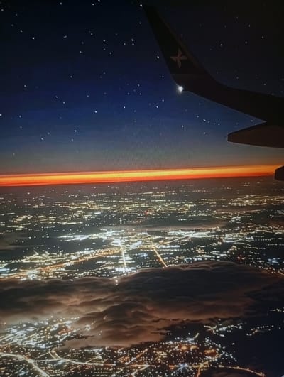 This image captures an aerial view of a city at twilight, likely Semey, Kazakhstan, from the perspective of an airplane window. The sky transitions from deep blue at the top, speckled with stars, to a vibrant band of orange and yellow along the horizon, indicative of sunset or sunrise. Below this celestial display lies the sprawling cityscape, illuminated by a dense network of lights. These lights form intricate patterns across the land, suggesting urban development and infrastructure.
Clouds, tinged with the warm hues of the sky, drift above parts of the city, casting shadows and adding depth to the scene. The wing of the airplane is visible in the upper right corner, a dark silhouette against the twilight sky. Its tail features a logo with a stylized star or cross. The overall impression is one of vastness and quiet observation, as the plane glides over a sleeping or awakening urban landscape. The image does not contain any discernible individuals or specific interactions, focusing instead on the relationship between the aerial viewpoint, the atmospheric conditions, and the urban environment.