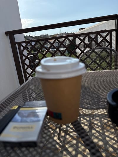 A close-up, eye-level, medium shot shows a disposable coffee cup with a white lid sitting on a textured metal table. To the left of the coffee cup, a pack of cigarettes is visible. In the background, a metal railing with a diamond pattern overlooks a residential area with white buildings and lush greenery under a clear blue sky. The lighting suggests it is daytime, with sunlight casting shadows across the table. The scene is likely on a balcony or patio in Chersonisos, Greece, given the architectural style of the buildings and the sunny weather.
