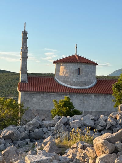 The image displays a stone church with a red-tiled roof, featuring a prominent bell tower or minaret on the left side and a dome-shaped section with a cross on top. The building is constructed with large, gray stone blocks, and a small, arched window is visible on the domed portion. The church is situated in a rocky, arid landscape with sparse green vegetation and trees. In the foreground, a dense pile of large, gray rocks creates a natural barrier. The background shows rolling, green hills under a clear blue sky with a few wispy clouds. The lighting suggests it is either early morning or late afternoon, with sunlight casting shadows on the building and highlighting the textures of the rocks and vegetation. The scene appears serene and isolated, with no people or animals visible. The location is identified as the city of Kovačko Polje, Montenegro.