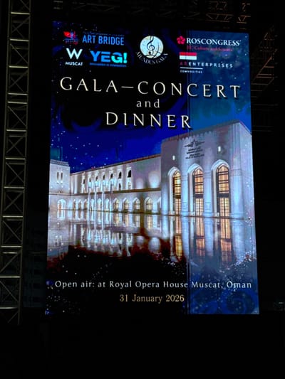 The image displays a large, vertically oriented digital screen acting as an advertisement or event announcement, likely positioned outdoors at night. The screen's content is a poster for a "GALA-CONCERT and DINNER."
The background of the poster features a dark, deep blue sky, transitioning to a lighter blue or perhaps reflecting water at the bottom. A grand, classical-style building, implied to be the Royal Opera House Muscat, is prominently displayed, illuminated with warm light spilling from its numerous arched windows. A shimmering reflection of the building is visible below it, suggesting water or a highly reflective surface. Small white specks are scattered across the darker parts of the background, possibly representing stars or a subtle artistic effect.
The main text, "GALA-CONCERT and DINNER," is centrally located, oriented vertically, and presented in a large, elegant serif font. Below this, horizontally aligned text reads: "Open air: at Royal Opera House Muscat, Oman" and "31 January 2026," clearly stating the location, setting, and date of the event.
On the left side of the poster, several logos and sponsor names are visible, also vertically oriented. From top to bottom, these include:
* "ROSCONGRESS" with "RC Culture and Science" below it.
* "ARENTERPRISES" with "COMMODITIES" below it, accompanied by a horizontal striped flag-like graphic (red, white, and green).
* A circular logo featuring a musical note and the words "MUNDUS GALA."
* "ART BRIDGE" with a stylized crown-like logo.
* Large text "W YEG!" followed by "MUSCAT" and "Innovation in interaction."
The screen itself is set against a completely dark environment, suggesting nighttime. Below the screen, a metal truss structure is visible, indicating that the screen is part of a larger outdoor setup, possibly a stage or a temporary installation for an event. The entire image is slightly tilted, giving a dynamic perspective of the large display.
