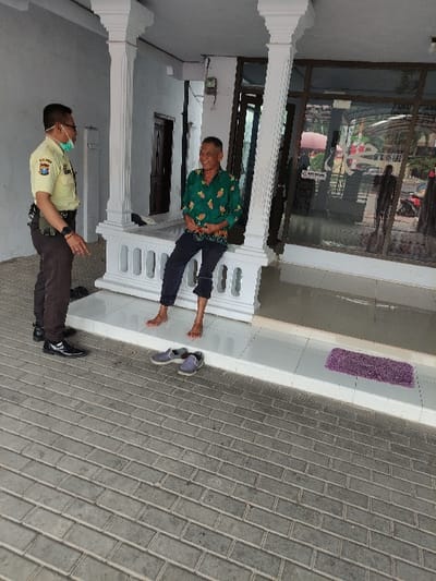 This image, rotated 90 degrees counter-clockwise, depicts two men interacting on what appears to be a paved outdoor area or porch in front of a building. The setting is likely in Situbondo, Indonesia, given the context.

In the foreground, an older man is seated casually on a low, white, ornate balustrade or bench. He is barefoot, wearing a green patterned short-sleeved shirt and dark trousers. He appears relaxed, with a slight smile or engaged expression, facing the other individual. Nearby, on the lighter tiled ground, are a pair of grey slip-on shoes and a pair of dark lace-up shoes, along with a purple mat.

Standing opposite the seated man is another man, dressed in a uniform consisting of a light-colored (possibly khaki) short-sleeved shirt, dark trousers, and dark polished shoes. He wears glasses and a light-colored face mask. A badge or emblem is visible on his left sleeve, typical of official or civil service attire in Indonesia. He also wears a watch on his left wrist. His posture suggests a professional interaction or conversation with the seated man.

The background features a building with large glass windows or doors, reflecting parts of the surroundings. White architectural pillars and a matching balustrade frame the scene. The ground is paved with grey rectangular tiles laid in a brick-like pattern, transitioning to a lighter-colored tiled surface closer to the building. The scene is illuminated by natural daylight, indicating it is daytime. No discernible text is visible in the image.