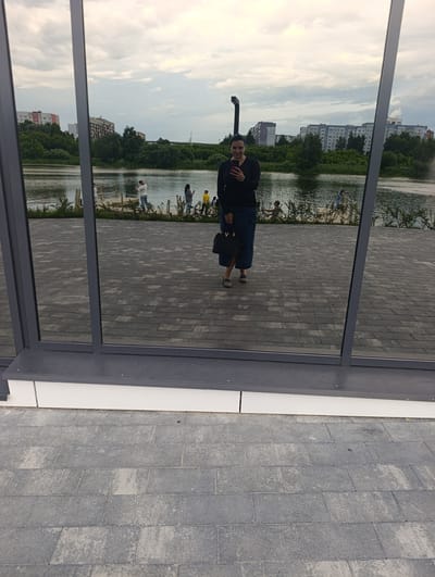 A full shot captured from a low angle shows a woman standing in front of a large window that reflects a city park with a lake. The woman is wearing a black long-sleeved shirt and a dark blue skirt, holding a black purse. She is looking at her phone, which is visible in her hand. The reflection in the window shows a calm lake with trees and buildings in the background. Several people are visible in the reflection, some standing near the lake and others on a wooden pier. The ground in front of the window is paved with gray rectangular tiles. The scene appears to be a public area in Penza, Russia.
