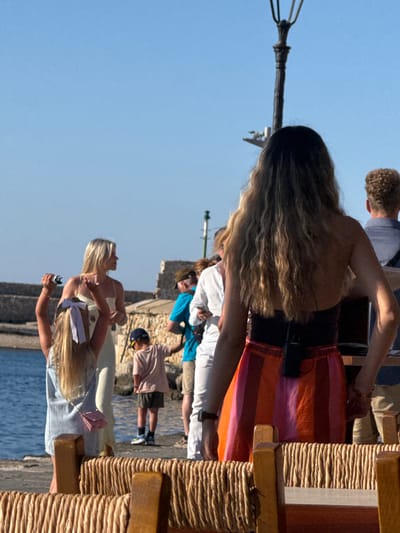 In Chania, Greece, a group of people are gathered outdoors, likely during a daytime event given the bright sunlight. The scene appears to be near a waterfront, with a body of water visible and a stone structure in the background, suggesting a coastal or harbor setting.
The focus is on several individuals, predominantly women, some of whom appear to be performing or posing. One woman in the foreground has long blonde hair and is wearing a light-colored top and skirt, holding a microphone or a similar object. She is gesturing with her hands, possibly singing or speaking. Behind her, another woman with blonde hair is also dressed in a light outfit, her hair partially obscuring her face.
Further back, a woman with long, wavy brown hair is prominently featured. She is wearing a dark top and a colorful striped skirt in shades of orange and pink. Her posture suggests she might be dancing or engaged in a spirited activity. There are other people in the background, including a man with curly hair and a person wearing a blue shirt. A young boy in a pink shirt and a baseball cap is also visible.
The setting includes wooden structures that resemble outdoor furniture or part of a stage, wrapped with rope. The overall atmosphere seems festive, with people enjoying the sunny weather by the water.