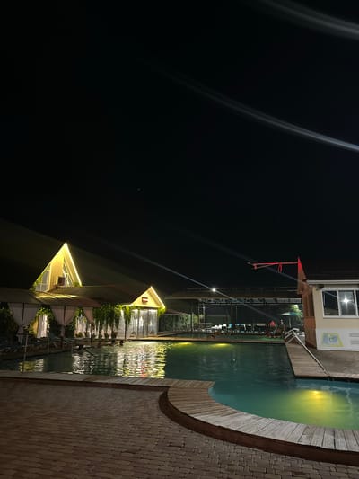This is a nighttime photograph of an outdoor swimming pool area, likely in Yaroslavskaya, Russia, given the context provided. The scene is illuminated by artificial lighting, casting yellow glows on the water and surrounding structures.
In the foreground, a brick-paved area leads to a wooden deck surrounding a kidney-shaped swimming pool. The water is a vibrant turquoise color, with reflections of lights shimmering on its surface. Several people are visible in the pool, some appearing to be swimming.
Along the edge of the pool, there are covered seating areas with white curtains and lush green hanging plants, suggesting a resort or restaurant setting. Beyond these, in the mid-ground, is a larger building with a distinctive triangular roofline, outlined with yellow lights. Further in the background, more structures and seating areas are visible under a canopy. The sky above is dark, with streaks of light, possibly from streetlights or other distant illumination. A red object with a loop hangs from a cantilevered arm extending from one of the buildings.