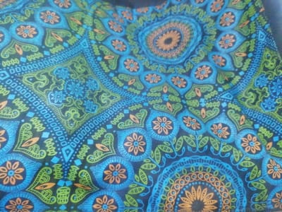 The image displays a close-up of a textile, specifically a fabric featuring an intricate, symmetrical pattern. The primary colors of the pattern are shades of blue (light blue, medium blue, dark blue), green (lime green, dark green), and orange, set against a dark blue or black background. The pattern includes mandala-like circular motifs, stylized floral elements with five petals, teardrop shapes, and various geometric designs such as squares, diamonds, and dotted lines, all arranged in a repeating, interlocking configuration. The fabric exhibits slight creasing, indicating it is not perfectly flat. There are no individuals, objects other than the fabric, or discernible actions present in the scene. The environmental context is limited to the fabric itself, offering no visual cues to suggest an outdoor or specific indoor setting beyond the close-up of the material. The location in the city of Johannesburg, South Africa, cannot be determined from the visual evidence provided.