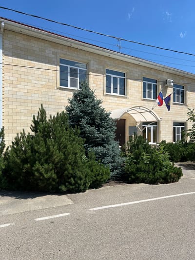 The image shows a two-story building with a light-colored brick facade, located on a sunny day in Takhtamukai, Russia. The building has several windows with white frames and a central entrance covered by a transparent awning. Two flags are visible near the entrance, one with red, white, and blue stripes and another with a blue background and a crest. In front of the building, there is a variety of coniferous trees and bushes. A paved road with white lane markings runs in the foreground. Overhead, power lines are visible against a clear blue sky. An air conditioning unit is mounted on the building's facade.