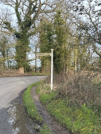 The image displays a rural road junction in North Norfolk, United Kingdom. A weathered white wooden fingerpost sign, topped with a spherical finial, stands on the right side of the frame. Its single visible arm points left, inscribed with the black capitalized word "CHURCH." To the left of the sign, a narrow, muddy dirt path runs parallel to an asphalt road that curves sharply to the left in the foreground. A significant puddle of water occupies the immediate edge of the road, reflecting the overcast sky and surrounding trees. The background and right side of the scene are densely vegetated with numerous deciduous trees, many extensively covered in dark green ivy, alongside undergrowth comprising green grass, dried brush, and brambles. A small, indistinct blue sign is partially visible through the foliage in the mid-ground on the right. The sky overhead is light and overcast. No individuals, animals, or vehicles are present.
