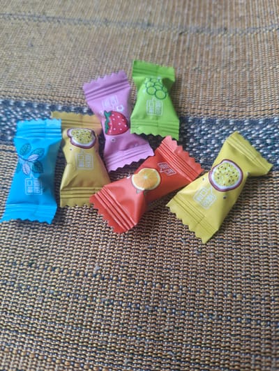 A pile of individually wrapped candies sits on a woven, textured surface, likely a placemat or tablecloth. The candies are in various bright colors: yellow, green, pink, red, and blue. Each wrapper features a simple illustration and Chinese text.

The yellow wrappers depict a slice of passionfruit. The green wrapper shows a cluster of grapes. The pink wrapper has a drawing of a strawberry. The red wrapper displays a slice of orange. The blue wrapper features a sprig of mint leaves. The Chinese text on each wrapper appears to be the same, likely indicating "flavor."

The lighting suggests daytime, possibly indoors. The overall impression is a colorful collection of sweet treats. No people are visible, and the activity is simply the presentation of the candies. The location context of ДТС «Чишки», Russia, is noted, but no specific visual cues in the image itself directly indicate this location.