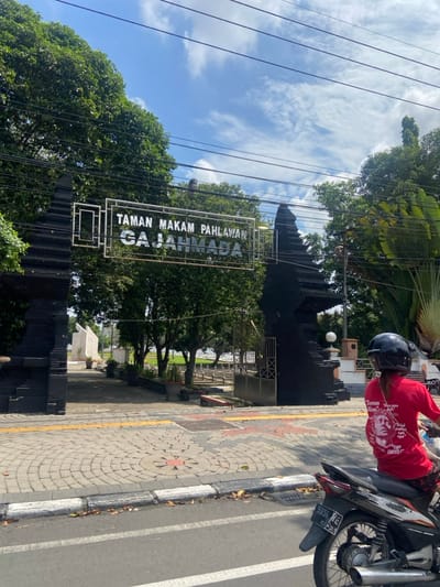 The image shows an entrance to a park in Mojokerto, Indonesia, located at coordinates -7.476, 112.44. The entrance is marked by two large black pillars with an archway between them. Above the archway is a silver sign that reads “Taman Makam Pahlawan Gajahmada.” The sign is situated in a frame of iron bars. The park appears to be a cemetery or memorial area with a green lawn and trees visible beyond the entrance. A person in a red shirt and a helmet is riding a black motorcycle toward the entrance. The motorcycle is stopped in the road just outside the park’s entrance. The ground in the foreground is a paved road with a cobblestone walkway leading to the entrance. The road is lined with trees, and the sky is a clear blue with white clouds. The scene is bathed in bright sunlight.
