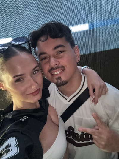 In this close-up selfie, a man and a woman are positioned centrally against a textured, dark background with bright, horizontal light streaks. The woman, on the left, has dark hair pulled back, revealing sunglasses perched on her head. She wears a black top with a large, light-colored 'B' graphic. The man, on the right, has dark, curly hair and a goatee. He wears a white t-shirt with dark trim and a black strap across his chest, and a watch on his left wrist. His right arm is around the woman's shoulders, and his right hand is raised, forming a peace sign. The woman's left arm is visible, resting on the man's chest. The overall scene suggests a casual outing or event. The image does not contain enough information to determine its location in Busmantsi, Bulgaria.