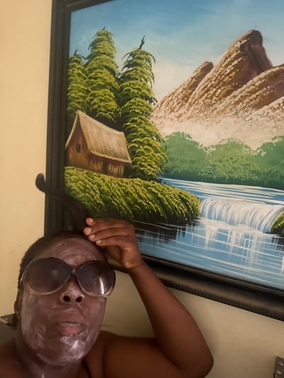 A person is lying on a light-colored surface, with an arm raised towards a painting hanging on a pale yellow wall. The person has sunglasses perched on their face, which is covered in a white substance, possibly a face mask. The painting, framed in black, depicts a landscape scene with what appears to be a waterfall, green vegetation, and rocky formations. The location is New Karu, Nigeria.