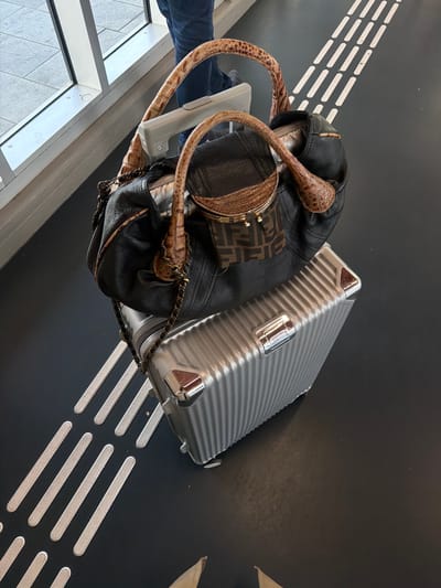 A high-angle shot shows a woman's luggage and a purse on a dark, textured floor with raised white lines, possibly a tactile paving surface. The luggage is a small, silver, ridged suitcase with metallic corners. Resting on top of the suitcase is a black leather handbag with distinctive patterned handles and a smaller brown pouch attached, featuring the Fendi logo. A person's legs clad in blue jeans are visible in the background, standing near a window with a tiled floor. The setting appears to be a public space, possibly an airport or train station, and based on the tactile paving, it is likely a location within the city of Rotterdam, Netherlands, which commonly uses such markings for accessibility.