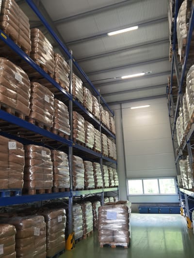 The image depicts the interior of a warehouse, specifically within the city of Lomets, Bulgaria. Tall, blue metal shelving units are densely packed with numerous stacks of goods, all individually wrapped in clear plastic. The goods appear to be sacks or bags, stacked on wooden pallets. The warehouse ceiling is high and features white corrugated metal panels with industrial fluorescent lighting fixtures illuminating the space. On the right side of the image, a large, white corrugated garage door or rolling shutter is visible, with a narrow horizontal window positioned above it, allowing some natural light to enter and revealing greenery outside. On the floor, a pallet of goods is positioned in the foreground, also wrapped in plastic. The floor itself is a light green, polished concrete surface showing some scuff marks and reflections. The overall impression is one of an organized and operational storage facility.
