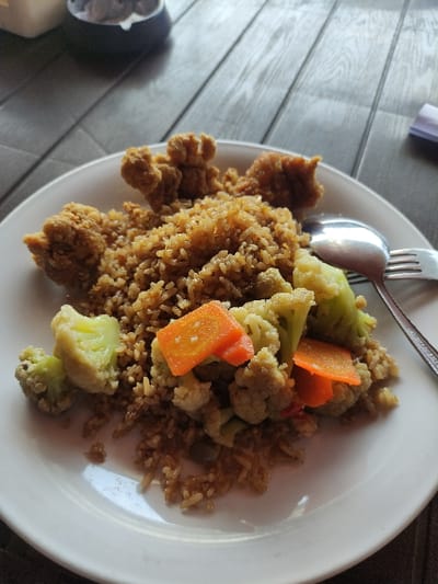 A white plate sits on a wooden table. The plate contains a serving of fried rice with fried chicken pieces, cauliflower, carrots, and other vegetables. A fork and a spoon lie to the side. The plate is partially obscured by a piece of fried chicken. The environment is a restaurant setting with a wooden table. The coordinates provided indicate a location in Gambiran, Indonesia.