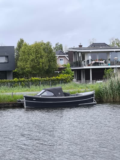 A black boat with a black canopy and silver trim is docked on a body of water. The boat is near a grassy shoreline with tall reeds. Behind the shoreline, there are houses and trees. The sky is overcast. The location is Oudorp, Netherlands.