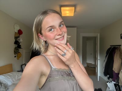 This image, reportedly taken in the city of Squamish, Canada, features a young woman smiling directly at the viewer within what appears to be a bedroom. The woman has shoulder-length blonde hair, fair skin, and visible freckles across her nose and cheeks. She has light-colored eyes, and her left ear is adorned with a large silver hoop earring and multiple smaller studs, while her right nostril displays a silver hoop nose ring. She wears a muted olive-green or light brown camisole with lace trim along the bustline, revealing visible natural hair in her left armpit. Her attire also includes a silver necklace with a cross-shaped pendant and several silver rings on her fingers, with her right hand gently resting against her chin.

The background shows light beige walls and a white ceiling featuring a square, frosted glass light fixture that emits a warm, yellowish glow. To the left, behind the woman, a bed with rumpled white bedding and a light wooden headboard is visible. Above the bed, a vertical arrangement of several hats in various colors, including white, red, brown, black, and yellow, hangs on the wall. To the right of the woman, a white interior door stands ajar, revealing a hallway with a wooden floor beyond. Adjacent to the door, a clothes rack is visible, laden with several garments, including dark, purple, and light brown items. A white tote bag partially printed with "BMW SERVICE" is discernible on the light-colored carpeted floor near the clothes rack.