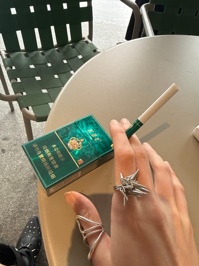 A person's hand is holding a green cigarette pack with a gold crown design and a single cigarette on a beige table. The hand is adorned with two silver rings. A woven green chair is visible next to the table, and part of a black shoe with studs is visible near the table leg. The text on the cigarette pack is in Chinese. The location is Hongkou District, China.