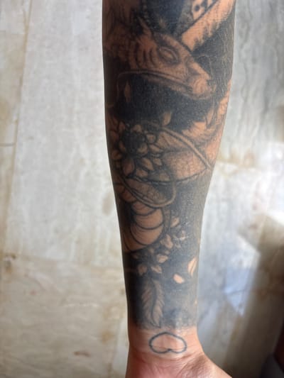 An arm, heavily tattooed with black ink, stretches across the frame. The tattoos include floral elements and abstract shapes. On the wrist, a small, simple heart outline is visible. The arm rests on a surface composed of light-colored, possibly marble, tiles.
The location of the photograph is unknown, and there is no reference to Cemagi, Indonesia.