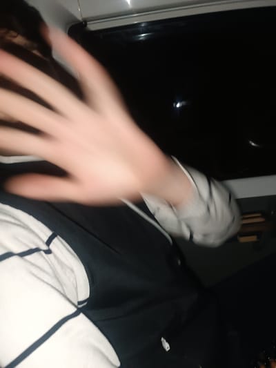 The image captures a candid, close-up shot of a person in a dimly lit setting, likely at night. The most prominent feature is a blurred hand, presumably the person's left hand, raised in the foreground with the palm facing the camera. This gesture suggests an attempt to block the camera or hide the face, creating a sense of spontaneity and motion blur.
Only a portion of the person's upper torso and arm are visible. They are wearing a long-sleeved shirt with distinct black and white horizontal stripes on the left side of the frame. Over this striped shirt, a dark, solid-colored garment, possibly a black or navy vest or top, is worn. On the right side of the image, another section of striped fabric, perhaps another sleeve or a piece of clothing, appears.
The background is largely dark and indistinct. A large, dark, reflective area occupies the upper right portion, resembling a window, possibly in a vehicle such as a car or bus, indicated by a faint light-colored frame or trim along its top edge. A small, bright, circular light reflection is visible within this dark background, hinting at external light sources or reflections from outside the window. The overall impression is one of a moment caught off guard, with the low light conditions and motion blur adding to the candid nature of the photograph. No text is visible in the image.