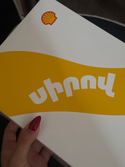A close-up image shows a partially visible white and yellow box bearing the word "Uruhru" written vertically in white on a yellow curved band. A red and yellow Shell logo appears on the white portion of the box. A portion of a hand with a red fingernail is seen holding the box. The environment consists of dark hues, possibly suggesting an indoor or shaded setting. The location and context of Zovuni, Armenia are not depicted in the image.