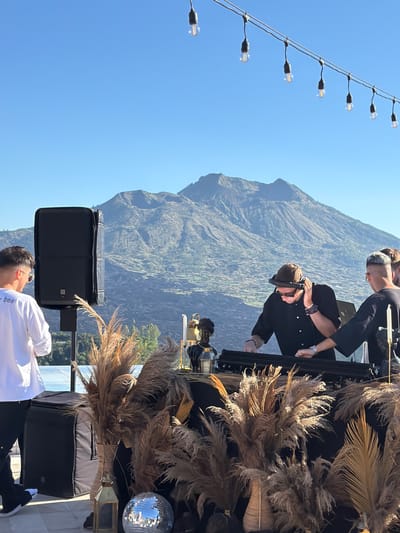 The image depicts an outdoor event setup against a backdrop of a mountain landscape, likely in the Kedisan area of Indonesia. In the foreground, there is a person wearing a white shirt. To the left, a string of light bulbs extends into the sky. In the center, two individuals are positioned at a DJ setup, with one person wearing headphones. The table is decorated with light brown, dried grasses. A disco ball is placed on the table. A tall, black speaker and case are visible. A mountain dominates the background with a clear blue sky overhead.