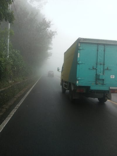 The scene depicts a foggy two-lane asphalt road in Siruppiddy, Sri Lanka. In the foreground, a box truck, predominantly teal-green on the rear and yellow on its visible side, occupies the right lane, moving away from the viewer. Its rear features two vertical securing bars and an obscured license plate area. A dark-colored passenger vehicle, possibly an SUV, is visible in the distant opposing lane, approaching the viewer, significantly obscured by heavy fog. The asphalt surface of the road appears wet. A continuous white line marks the left edge of the road, running parallel to a concrete drainage ditch and a dense wall of green vegetation, including bushes and trees, which lines the embankment. A slender utility pole or tree trunk is partially visible on the far left. The sky and distant landscape are completely obscured by the thick fog, limiting visibility. No individuals are visible.