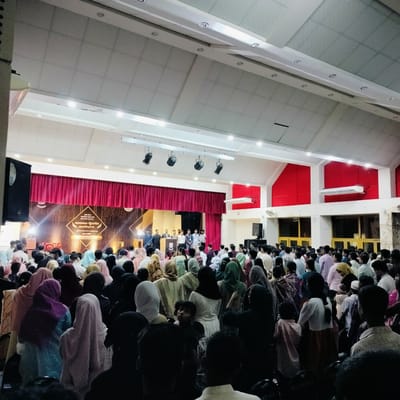 A densely populated auditorium in Lalmohan, Bangladesh is filled with attendees facing a stage. Onstage, a group of individuals stands behind a podium. The event appears formal, evidenced by the stage backdrop and sound equipment. The audience is diverse, with many women wearing head coverings. The hall has a high ceiling and bright lighting.