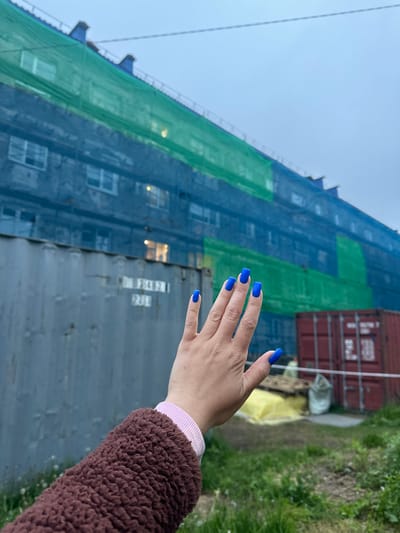 An outdoor medium shot captures a person's hand with bright blue fingernails extended towards a large building covered in blue and green netting. The netting obscures most of the building's facade, but glimpses of windows and lights are visible through the mesh. In the foreground, a corrugated metal shipping container with faded markings stands to the left of the hand. To the right, another red shipping container is partially visible, along with some construction materials, including a yellow tarp and a pallet. The background shows a slightly overcast sky and an area of overgrown grass. The scene appears to be an urban or industrial area, possibly undergoing construction or renovation, in Vilyuchinsk, Russia. The person is wearing a brown fuzzy jacket with a pink cuff visible at the wrist. The hand is positioned in a relaxed, open posture, with fingers slightly splayed.