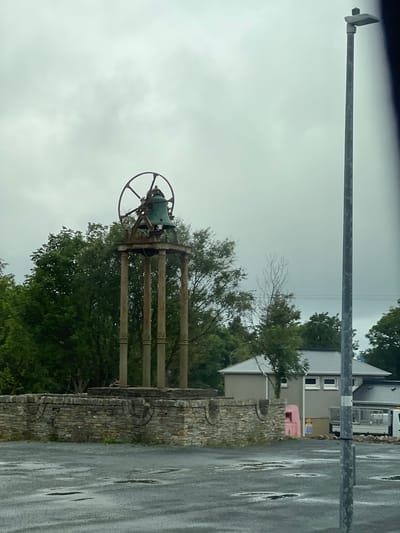 The image depicts an outdoor scene in Derrybeg, Ireland, under a cloudy sky. In the center, a large, old, greenish-bronze bell sits atop a rustic metal structure supported by four weathered wooden pillars. This entire assembly is erected on a low, circular stone wall. The ground in front of the wall is a dark, wet asphalt surface with several puddles, indicating recent rain. To the right of the bell structure, a modern grey lamppost stands prominently. In the background, there are trees, a grey-roofed building with white-framed windows, and a pink structure. Further to the right, a white truck is partially visible behind the lamppost. The overall atmosphere is overcast and damp.
