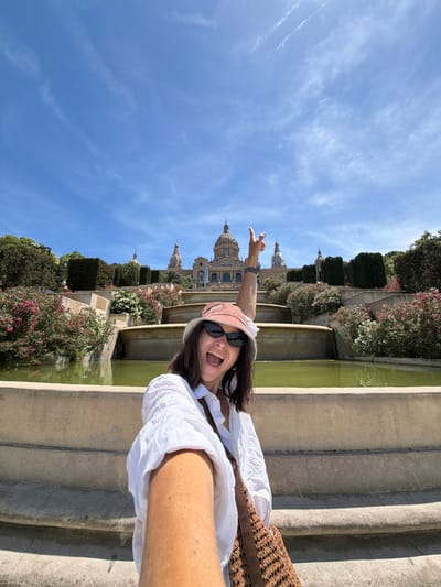 The image is a selfie taken by a woman in front of a grand building with domes, situated behind a tiered fountain and lush greenery. The woman is wearing a white shirt, sunglasses, and a pink hat. She is smiling and holding her right arm up with her index finger pointing towards the sky. The background shows a large, ornate building with multiple domes and columns, likely a museum or palace, with a clear blue sky above. The building's architecture suggests it might be a prominent landmark in a European city. The area appears to be a public space, possibly a park or a plaza, with a large fountain and well-maintained gardens. The scene is brightly lit by the sun, indicating a daytime setting. The woman's excited pose and the picturesque backdrop suggest she is on vacation or sightseeing. The location is the National Art Museum of Catalonia, located on Montjuïc hill in Barcelona, Spain, which is near L'Hospitalet de Llobregat.