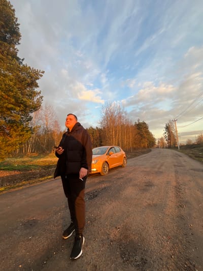 The image captures an outdoor scene at golden hour, characterized by warm, low-angle sunlight casting long shadows and illuminating clouds with soft orange and blue hues. The primary subject is a young man, standing on a dirt road and looking towards the viewer. He wears a black padded jacket, black trousers, and black sneakers with white soles. He holds a lit cigarette in his right hand and a mobile phone in his left.
Beside the man and slightly behind him is a bright orange hatchback car, parked on the unpaved road. The road surface is composed of dirt and small stones, suggesting a rural or semi-rural environment. The background features a landscape with various trees, including bare deciduous trees and green conifers, indicating either late autumn or early spring. Overhead, power lines are visible against the cloudy sky. The overall setting is consistent with an outdoor location in Voronino, Russia, with no discernible text visible in the frame.