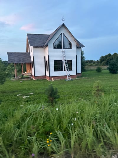 A white, two-story house, currently under construction, is situated on a grassy lot in Abushkovo, Russia. The house features dark-trimmed windows, a dark roof, and a prominent triangular window on the upper level. A silver ladder leans against the side of the house, extending to the roof. A covered porch with wooden pillars is attached to the left side of the house. The surrounding environment consists of green grass, scattered trees, and a vast, partly cloudy sky with hints of pink and blue. In the foreground, tall, green grass with some small wildflowers is visible.