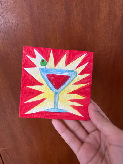 A person's hand holds a small, square painting of a martini glass with a green olive. The background of the painting is red with a yellow and white starburst pattern. The painting is placed on a wooden surface. No information is available about the location in the city of Maracay, Venezuela.
