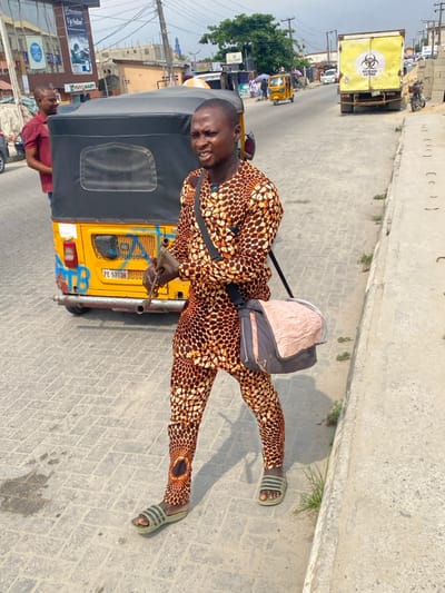 The image depicts a daytime street scene in Langbasa, Nigeria. In the foreground, a male with dark skin and short hair walks right-to-left across a light grey paved road. He wears a long-sleeved, collared two-piece outfit with a brown, black, and white mottled or spotted pattern, and green-striped slide sandals. He holds a metallic, pipe-like object in his right hand, pointed downwards. A black shoulder strap crosses his chest, supporting a grey and light pink/beige shoulder bag resting on his left hip. His head is turned slightly to his left, and his mouth is partially open.

Behind him, to the left, a yellow Keke Napep (auto-rickshaw) is parked, displaying a partial license plate "PE 576QN" and blue graffiti on its rear. A second male in a maroon t-shirt stands near the rear of the Keke Napep, looking towards the left.

In the mid-ground and background, the road continues, lined with various buildings and sparse vegetation. On the left, a multi-story building with reflective windows shows "Up Solar" and "Phy Smart" signage. Further down the street on the right, a large yellow box truck is parked, featuring a black biohazard symbol and "WASTECARE SYSTEMS" text on its rear. Additional distant vehicles, including other Keke Napeps, are visible along the road. The sky is overcast. A raised concrete curb separates the paved road from a narrow dirt and grass verge on the right.