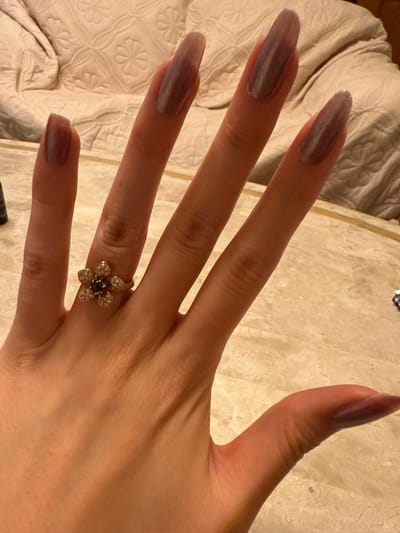 A dorsal view of a light-skinned left human hand is depicted, with fingers slightly spread and resting on a light-colored, veined surface within the City of Kawasaki, Japan. The fingernails, including the thumb, index, middle, ring, and pinky, are long and almond-shaped, with the pinky nail appearing shorter than the others. All visible nails are coated with a translucent, shimmering purplish-gray polish. A yellow-gold toned ring, fashioned as a flower with a central dark gemstone surrounded by smaller clear stones, is worn on the ring finger. In the blurred background, a light-colored textile item with a subtle patterned design, consistent with upholstered furniture or bedding, is visible. A dark, partially visible cylindrical object is located on the surface to the left of the pinky finger.