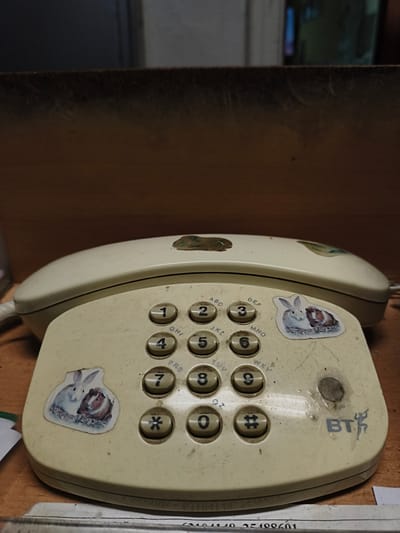 A close-up, eye-level shot captures a cream-colored, vintage BT corded telephone with a keypad on a wooden desk. The phone is adorned with two stickers: one of a rabbit and guinea pig on the left side of the keypad, and another similar sticker partially visible on the top left of the phone's handset. The keypad features numbers 0-9, asterisk, and hash symbols. A small, worn circular mark is present on the right side of the keypad, above the "BT" logo which is visible in dark gray. The phone and its surroundings appear dusty, suggesting it has not been recently cleaned. The background consists of a blurred wooden surface and a dark, out-of-focus area. The overall scene evokes a sense of disuse and the passage of time. There is no identifiable location information or people present in the image.