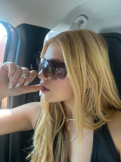 A fair-skinned woman with long, blonde hair wears sunglasses and a black top, positioned in a vehicle. She's touching her lips with a finger adorned with rings, one shaped like a flower. Her nails are polished. A delicate pearl necklace with multiple charms hangs around her neck. The car's interior, including the dashboard and a portion of the door, is visible in the background.