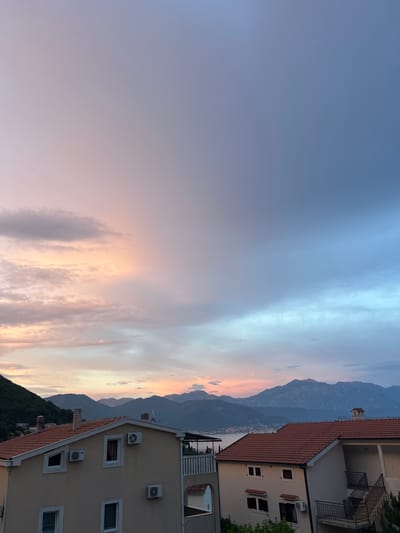 The image shows a picturesque sunset over a coastal town in Baošići, Montenegro. The sky is painted with soft hues of blue, pink, and orange, with wispy clouds stretching across the horizon. In the foreground, a multi-story building with a tiled roof is partially visible, featuring several windows and air conditioning units. To the left, lush green hills descend towards the sea, which is faintly visible in the distance. Towering mountains form a dramatic backdrop, their silhouettes softened by the fading light. The scene evokes a sense of tranquility and natural beauty, characteristic of a peaceful evening by the sea. The time of day is clearly sunset, indicated by the warm, diffused light and the colors in the sky. No people or text are visible in the image.