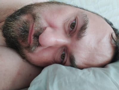 An extreme close-up shot shows a Caucasian man's face, lying on his side, with his head tilted. The man has brown hair and a beard with some gray strands. His eyes are open, and he appears to be looking towards the camera. The lighting is soft, and the background is a textured white surface, possibly a bedsheet. There are no other people or objects visible in the image. The image does not contain any contextual information about the city of Konstantinovo, Russia.
